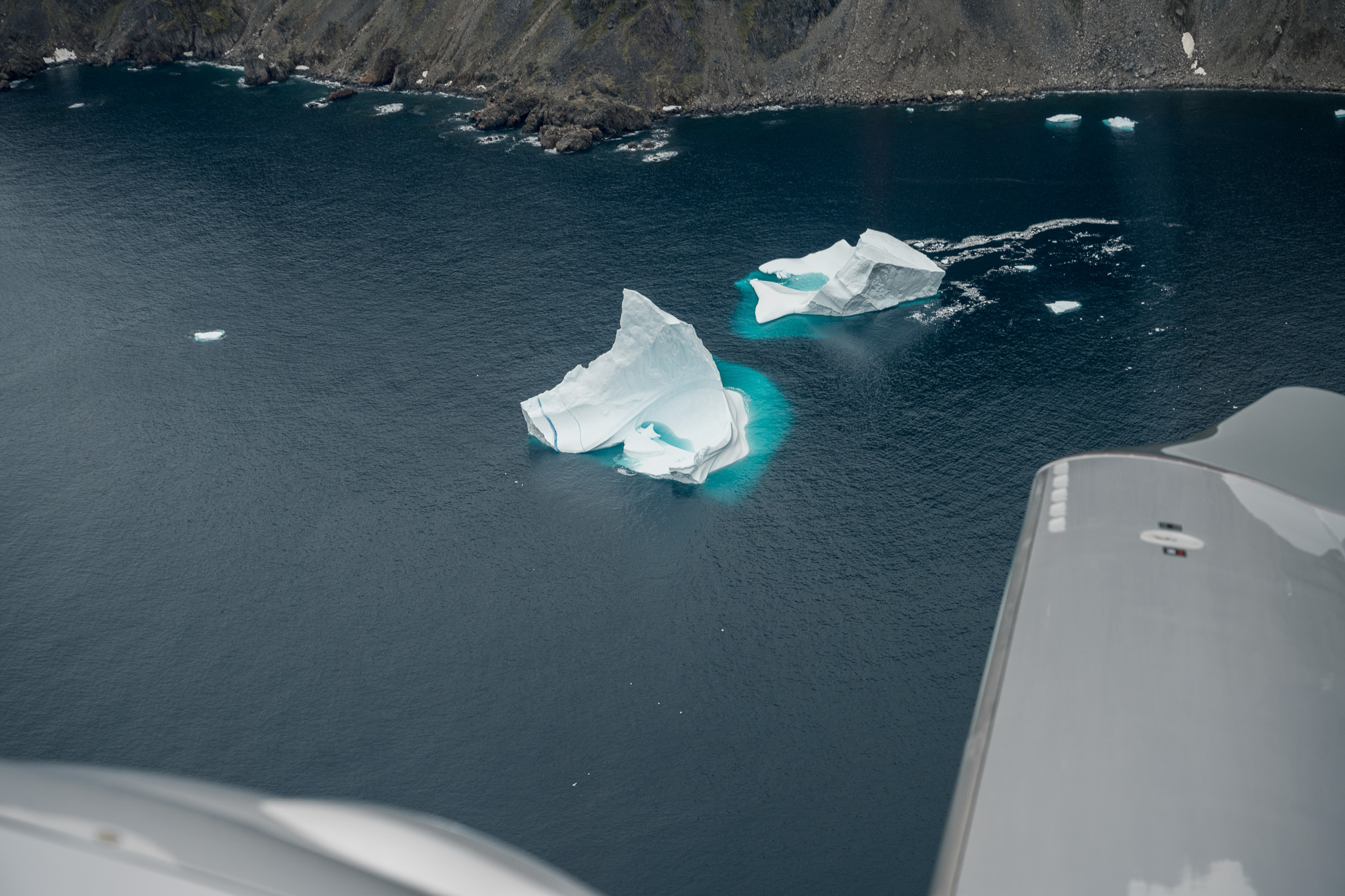 Two sculpted icebergs in dark Greenland water beside the aircraft wing